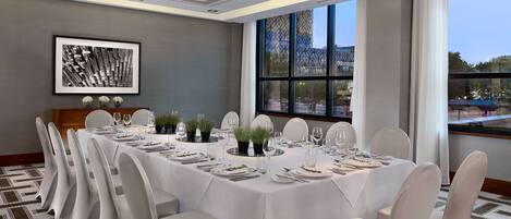 A conference room with a long table set for a meeting, large windows, and a framed artwork on the wall.