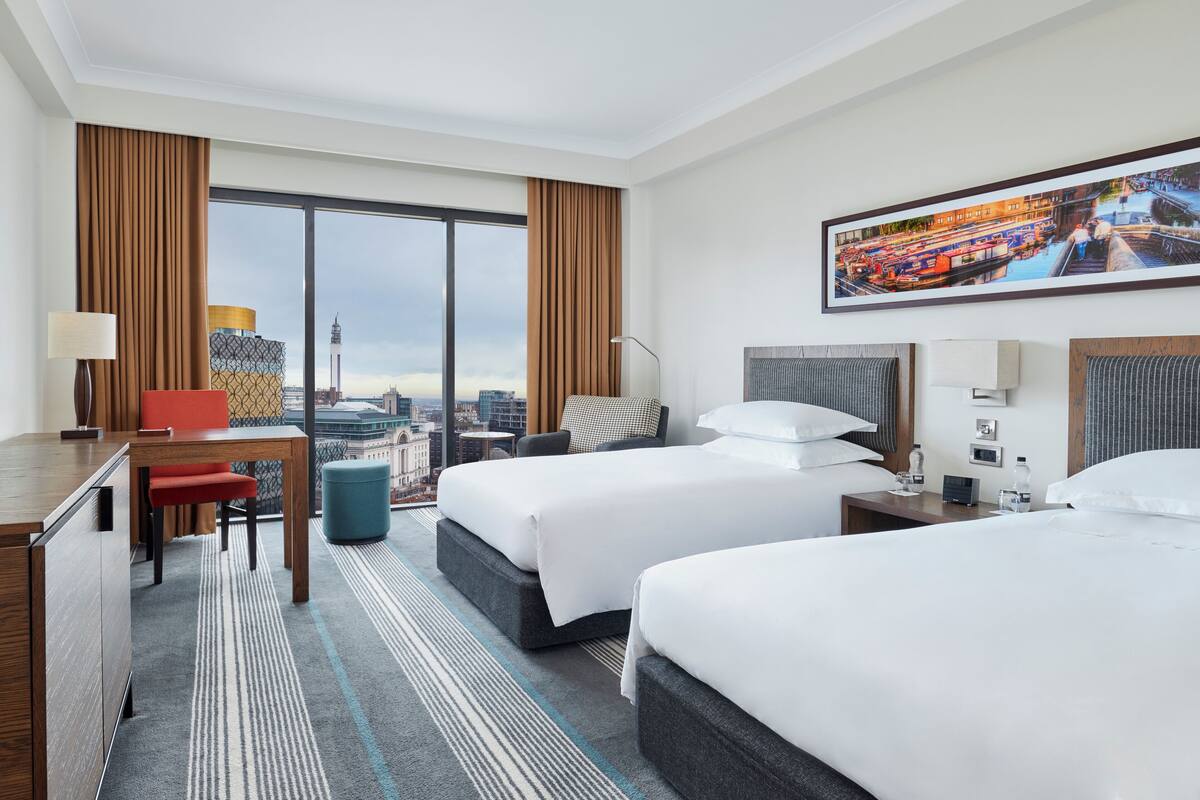 A hotel room with two beds, a desk, a chair, a small table, and a large window offering a city view.