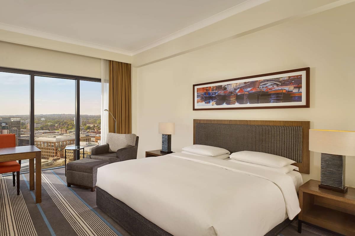 A hotel room with a large bed, a desk, a chair, and a view of the city through large windows.