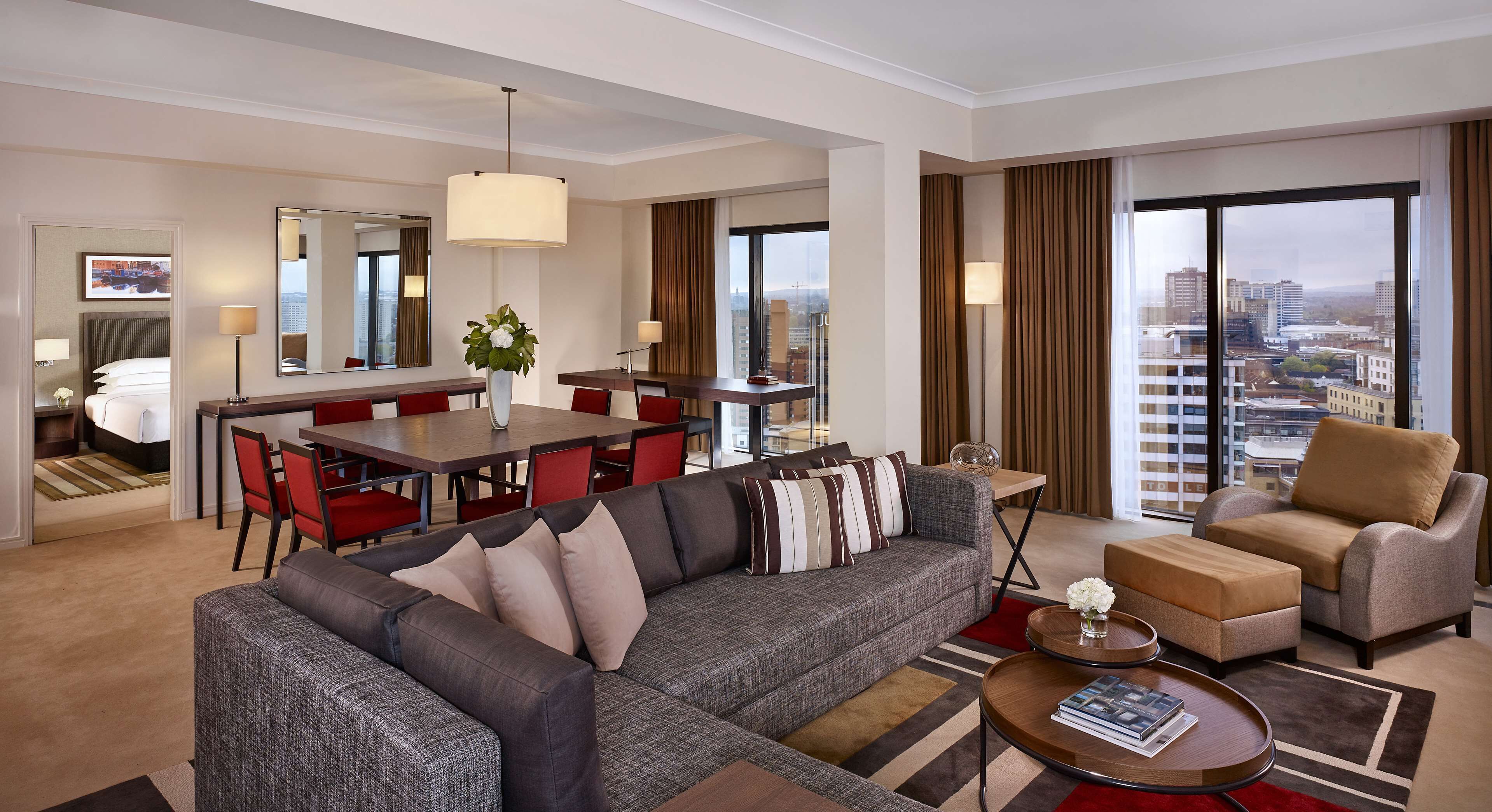 A modern hotel room with a gray sofa, red chairs, a dining table, and a large window with a city view.