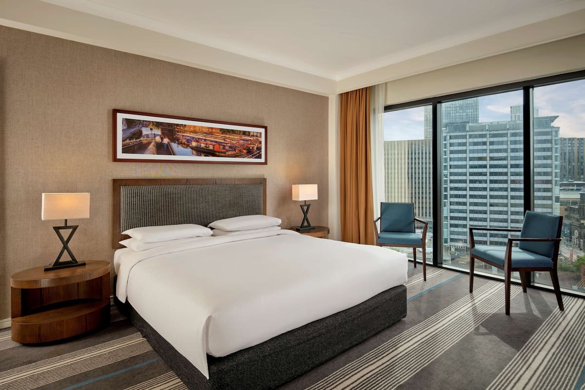 A hotel room with a large bed, two side tables with lamps, a framed cityscape picture on the wall, and a view of tall buildings through the window.