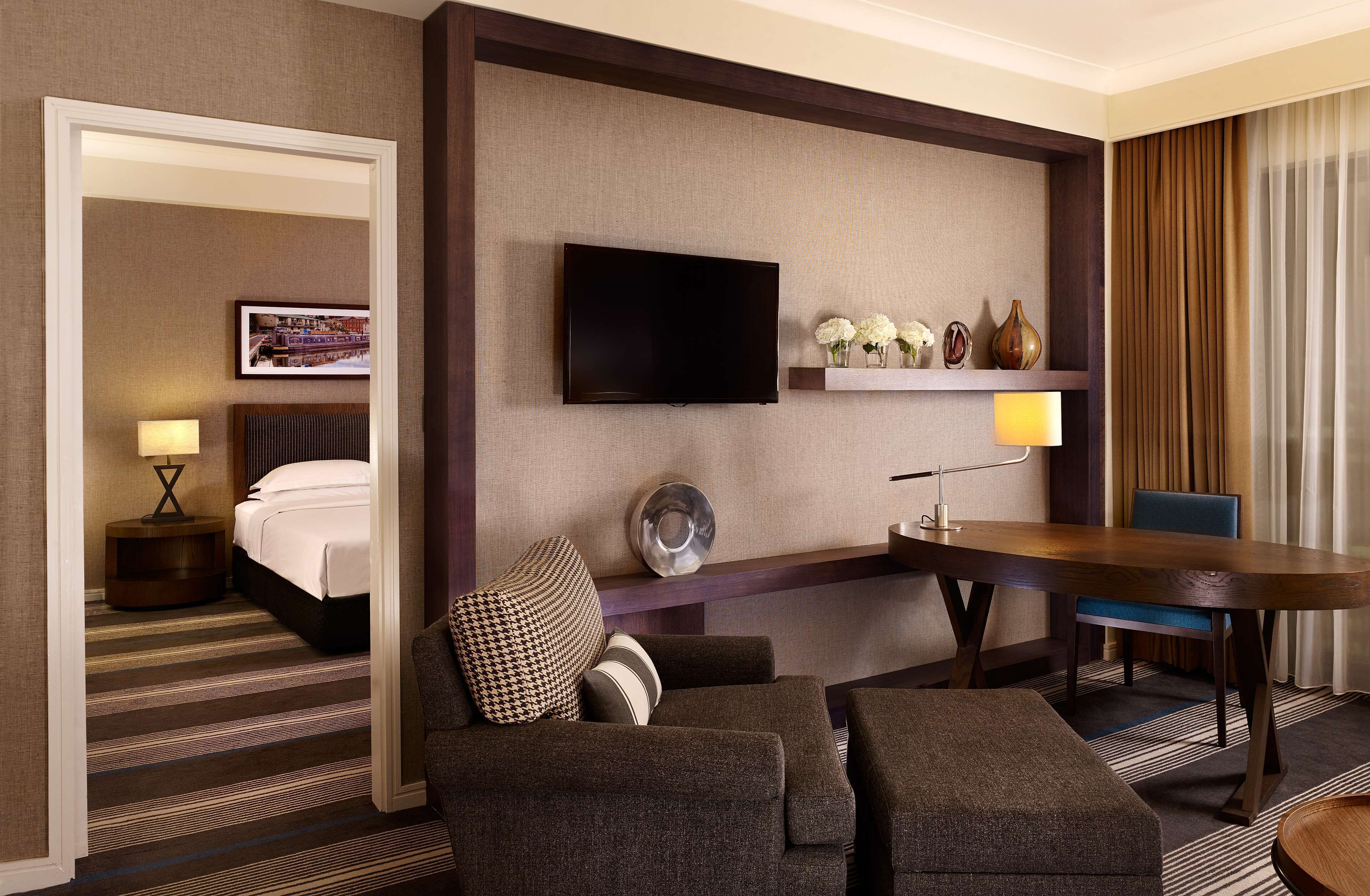 A hotel room with a bed, a TV mounted on the wall, a dining table, and a seating area with a sofa and chairs.