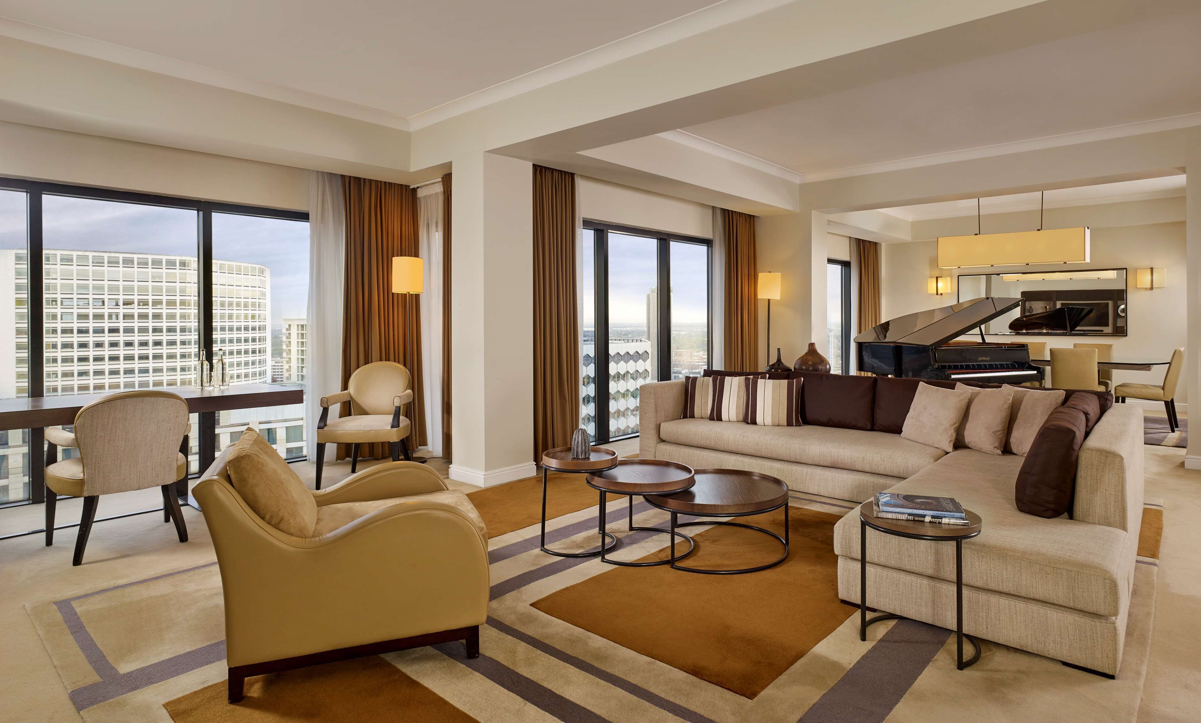 A spacious living room with a sofa, armchairs, and a coffee table. There is a piano and a dining area with a table and chairs. Large windows provide a view of the cityscape.