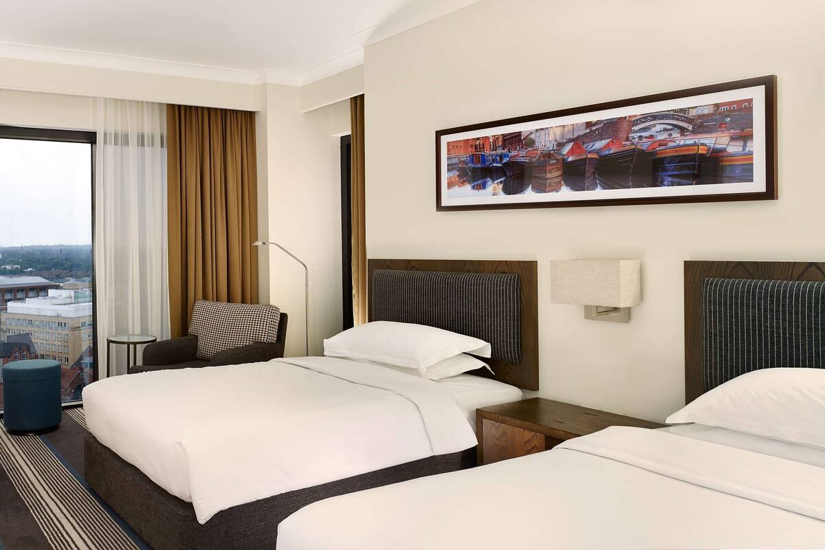 A hotel room with two beds, a dining table, a chair, a small round table, and a large window with curtains.