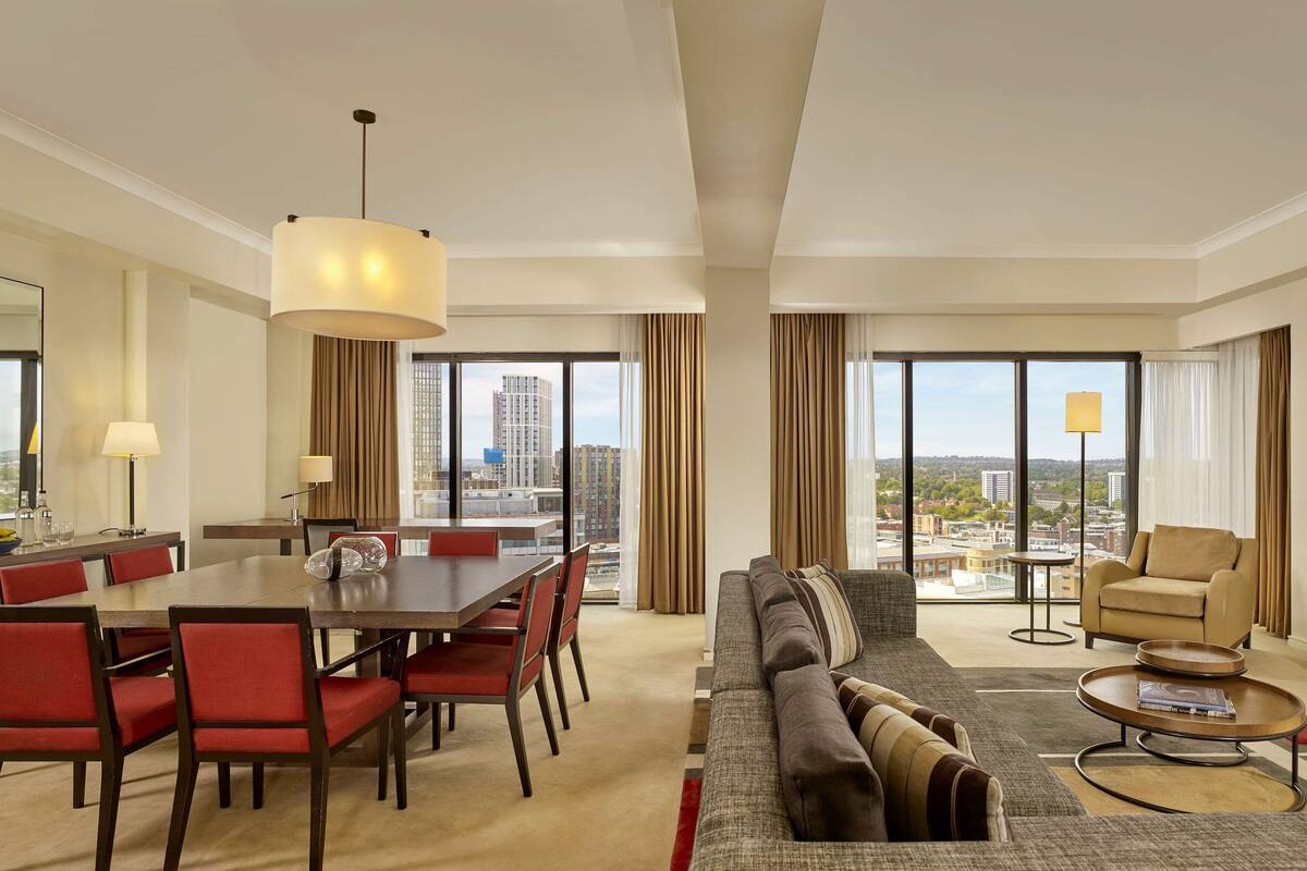 A spacious hotel room with a dining area, a sofa, a coffee table, and a view of the city.