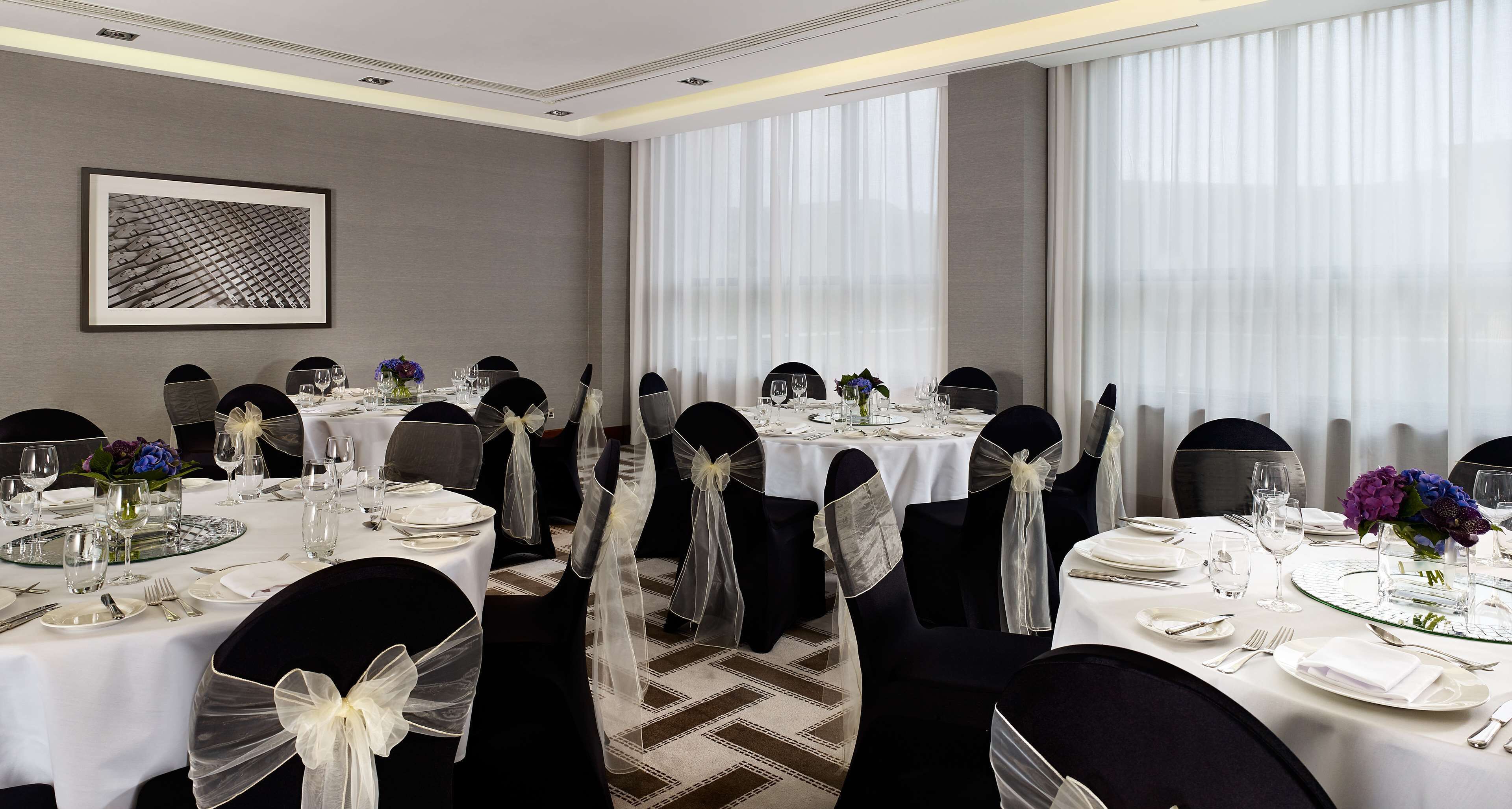 A banquet hall with round tables, black chairs with white sashes, and large windows with sheer curtains.