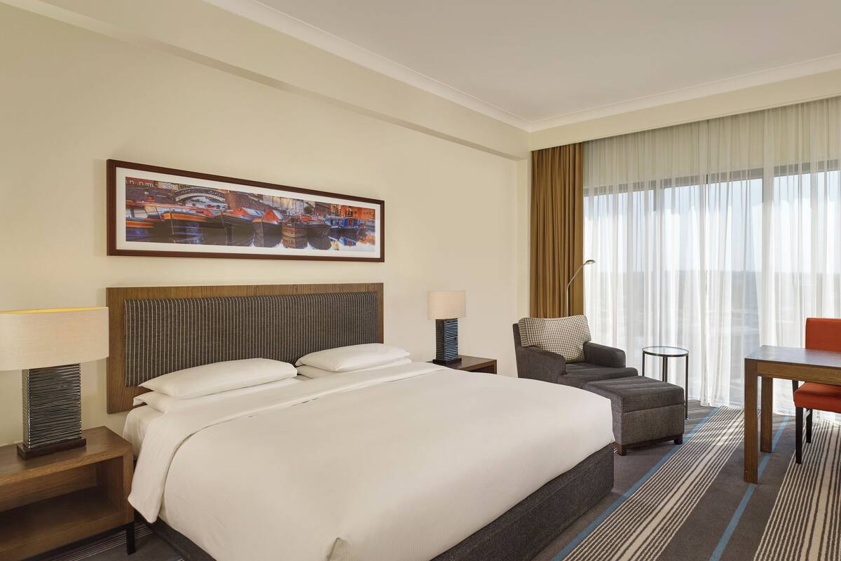 A hotel room with a large bed, a sofa, a desk, and a chair. There is a painting on the wall and a window with curtains.