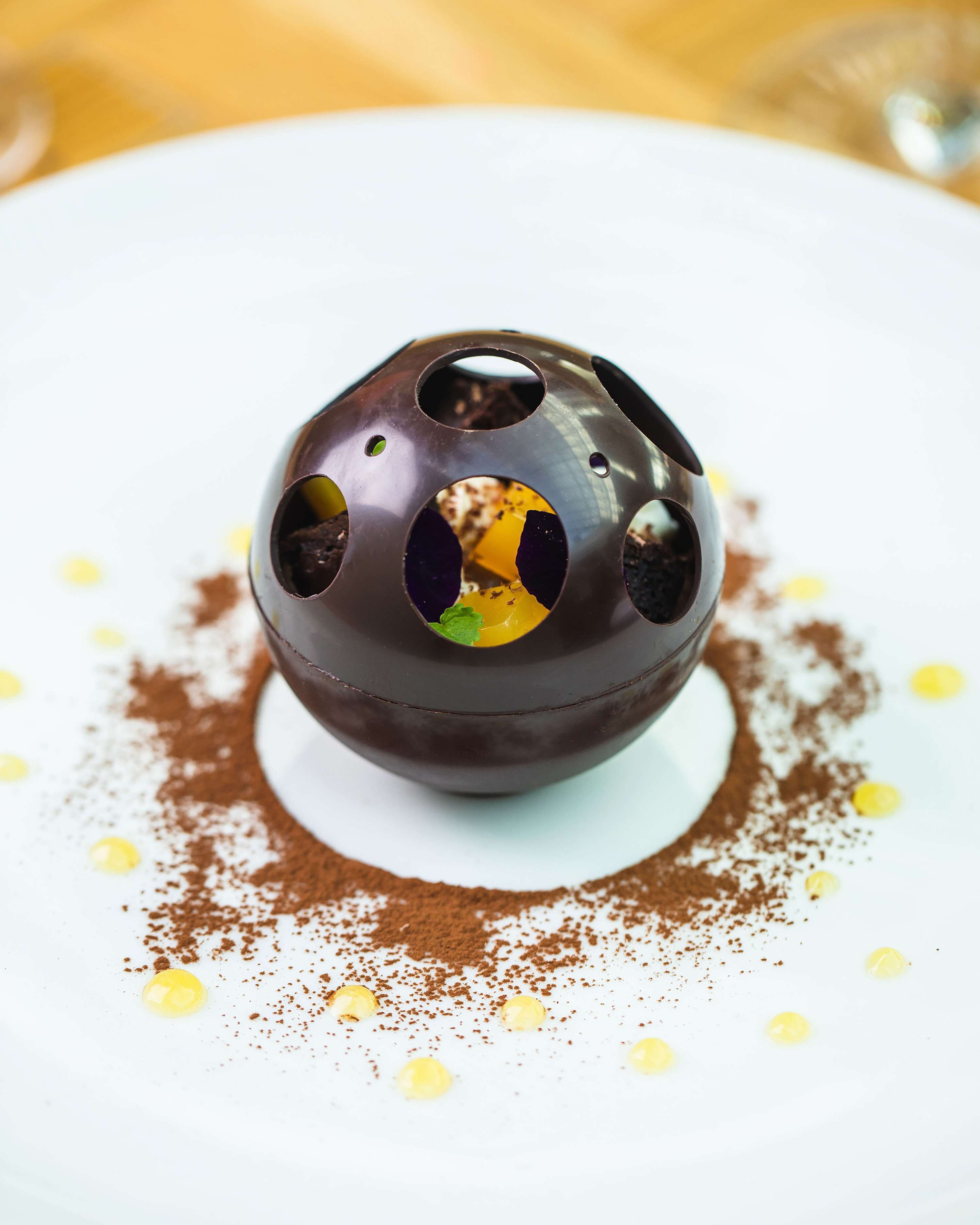 A chocolate dessert with a spherical shape and multiple holes, served on a white plate with cocoa powder and yellow dots.