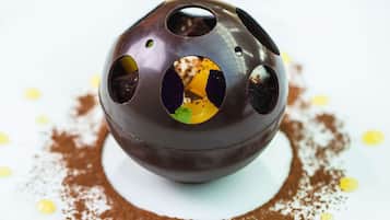 A chocolate dessert with a spherical shape and multiple holes, served on a white plate with cocoa powder and yellow dots.