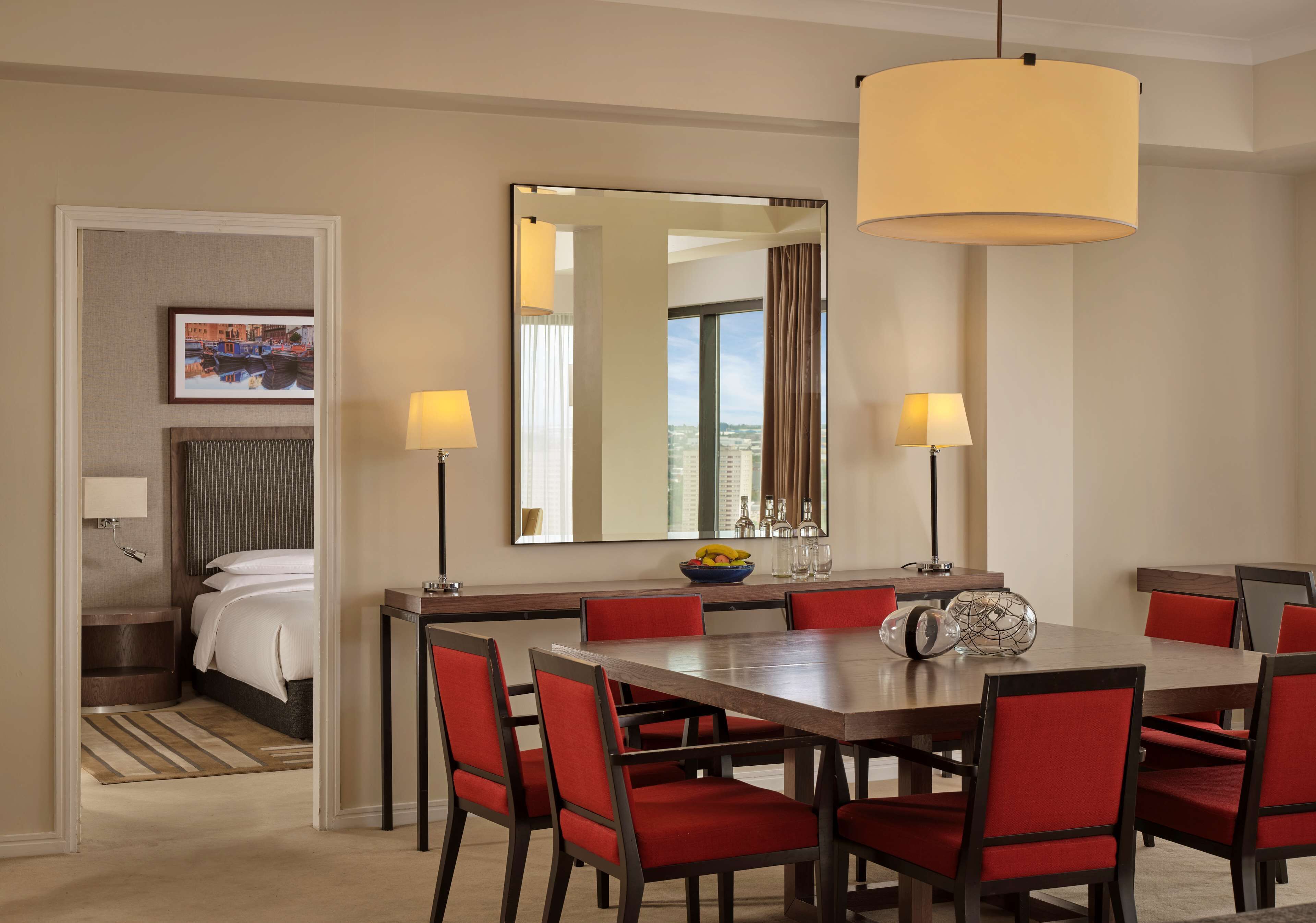 A hotel room with a dining area featuring a large table, red chairs, and a modern chandelier.