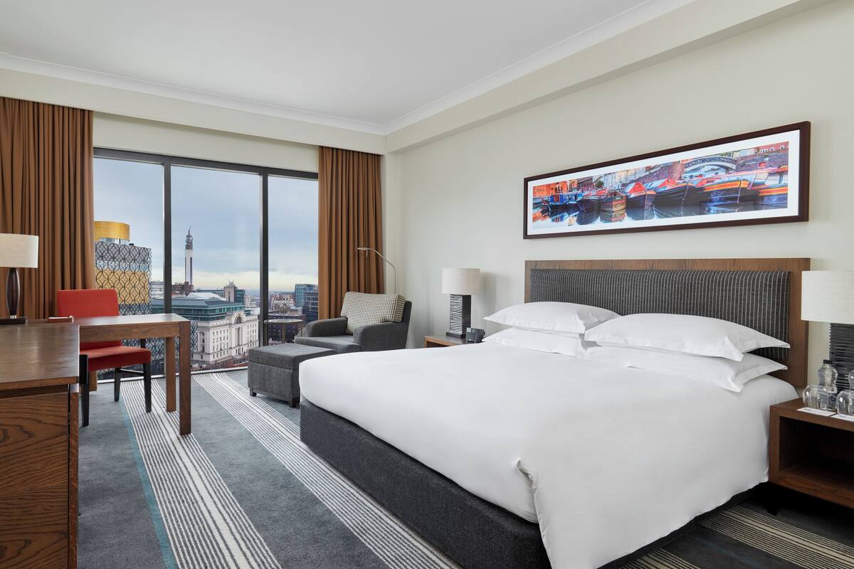 A hotel room with a large bed, a desk, a chair, a sofa, and a view of a cityscape through large windows.