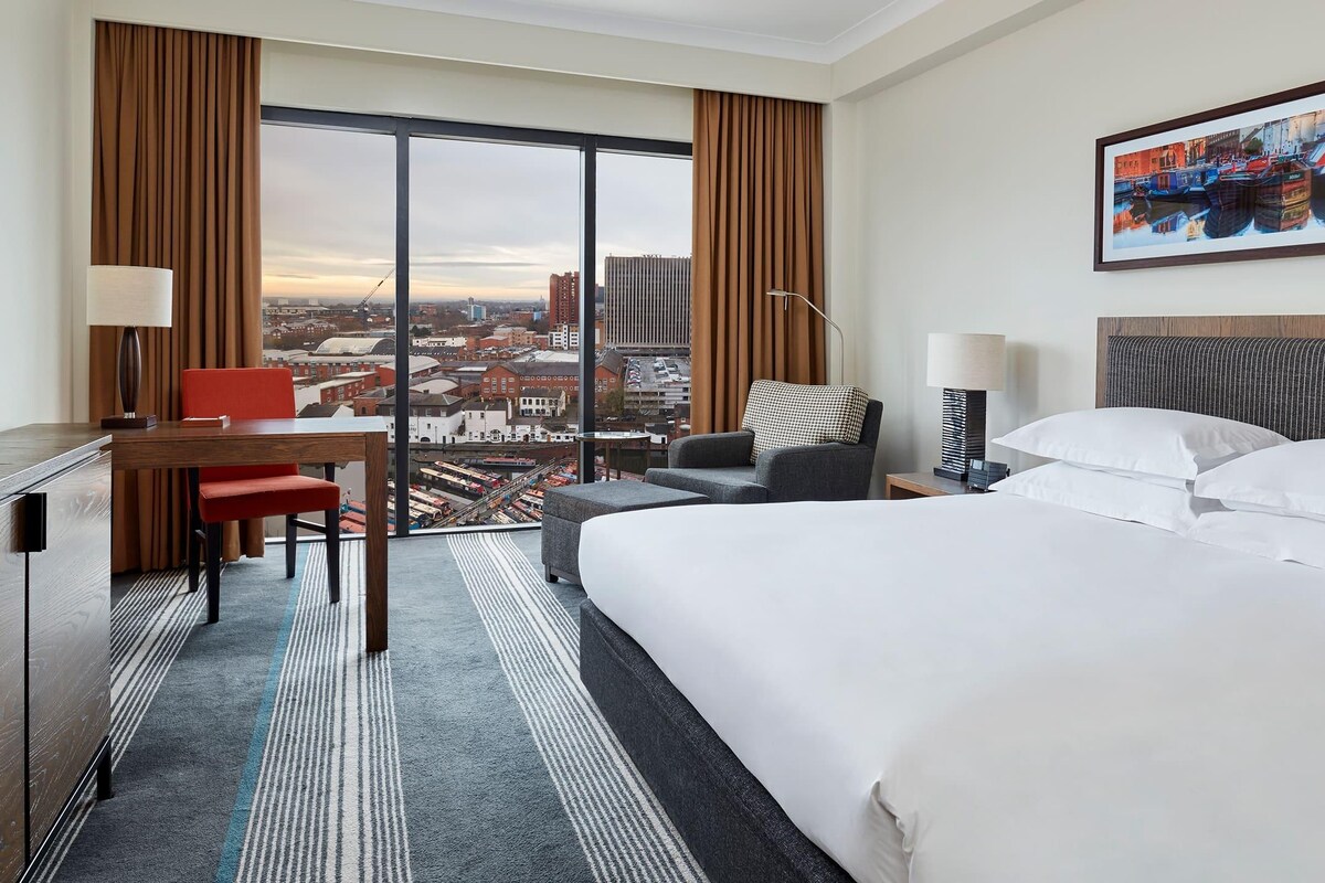 A hotel room with a large bed, a desk with a chair, a sofa, and a view of the city through large windows.