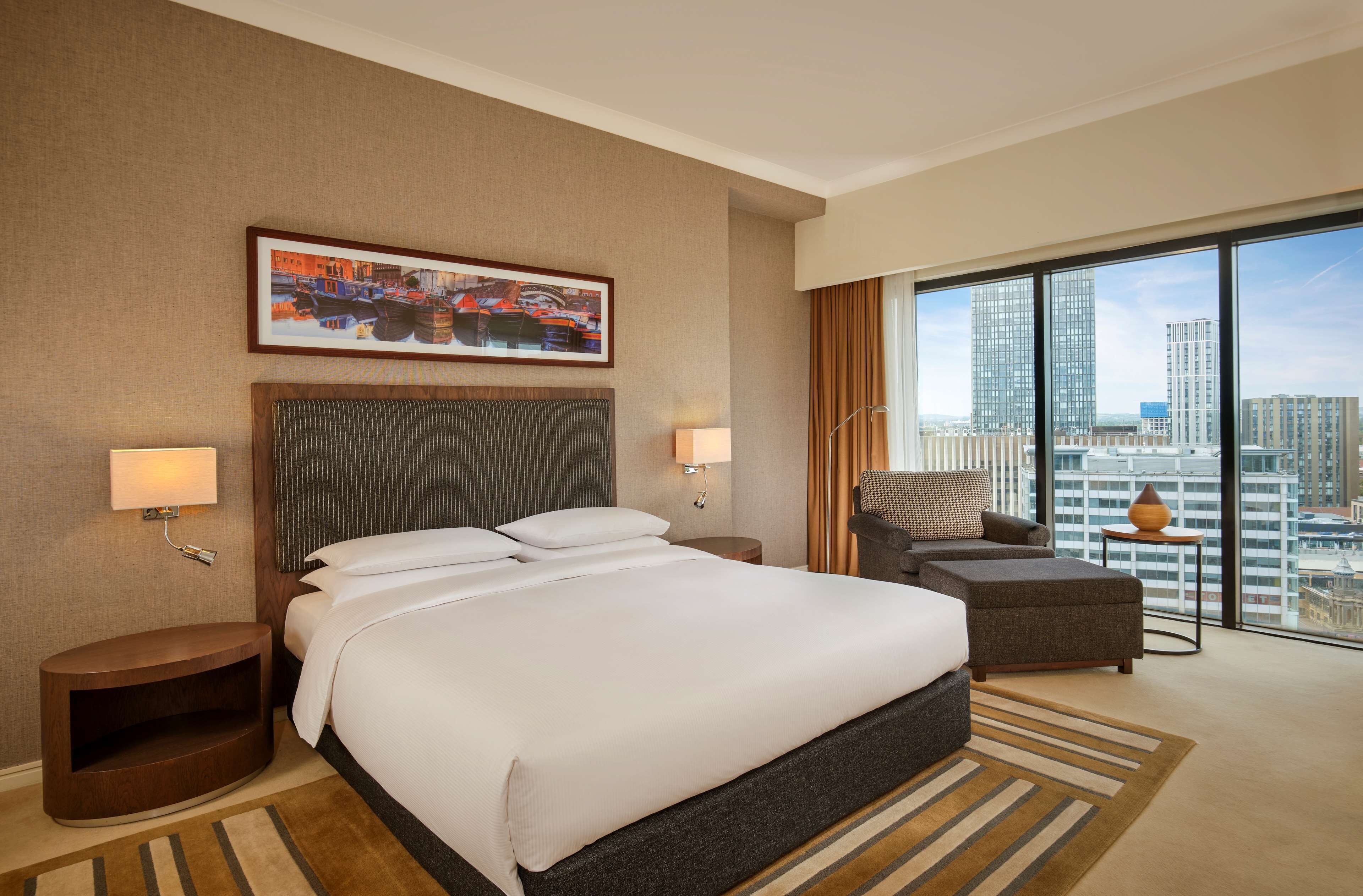 A hotel room with a large bed, two bedside tables, a chair, a view of the city through large windows, and a painting on the wall.