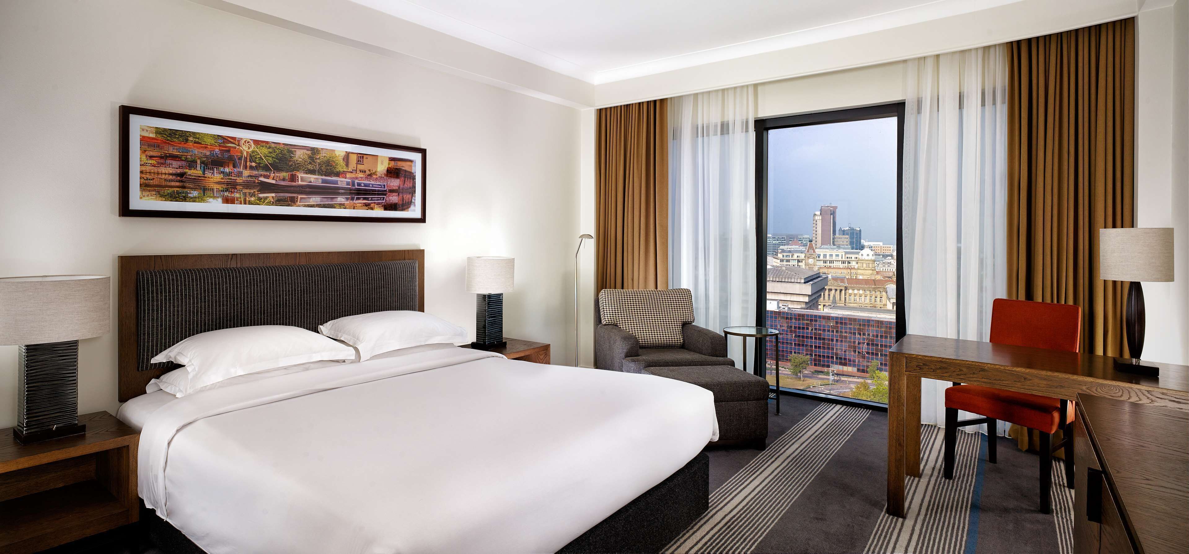 A hotel room with a large bed, a desk, a chair, a view of the city through a large window, and a painting on the wall.