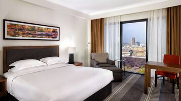 A hotel room with a large bed, a desk, a chair, a view of the city through a large window, and a painting on the wall.