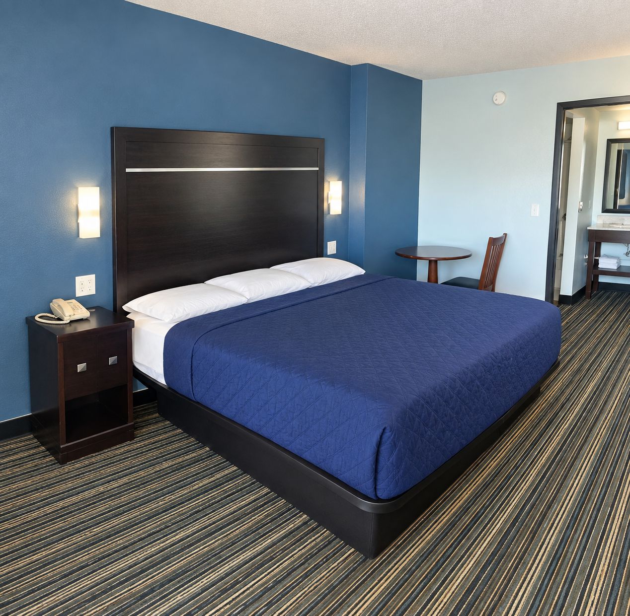 A hotel room with a large bed, a bedside table with a phone, a small table with a chair, and a bathroom visible through an open door.