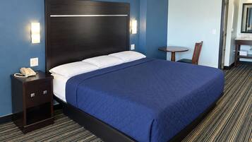 A hotel room with a large bed, a bedside table with a phone, a small table with a chair, and a bathroom visible through an open door.