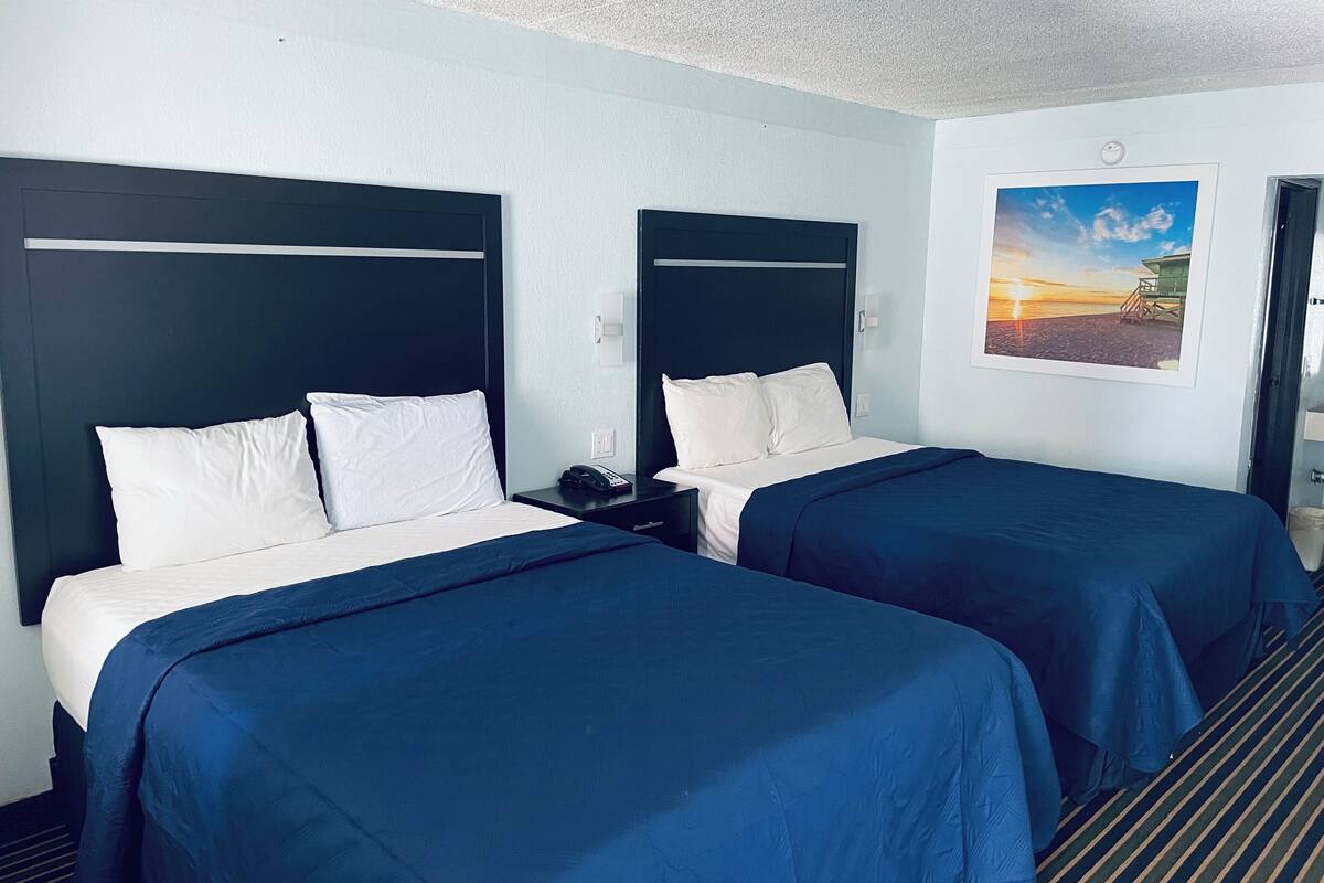 A hotel room with two beds, each with white linens and dark blue bedspreads. There is a nightstand with a phone and a wall-mounted light switch. A framed picture of a beach scene is on the wall.