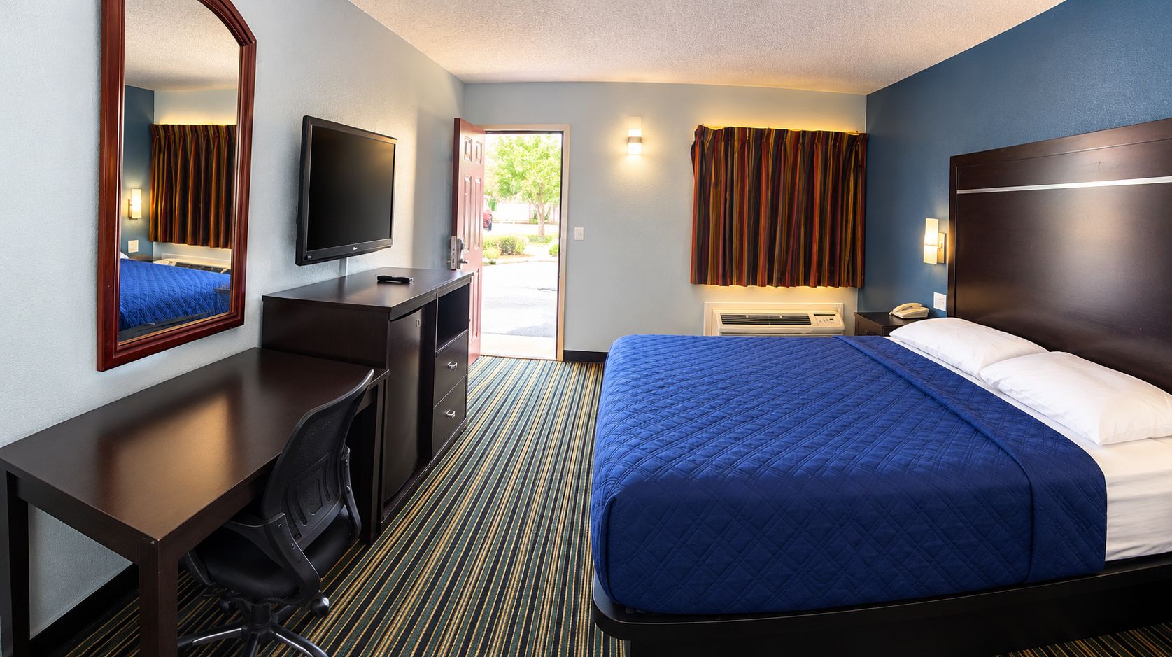 Habitación de hotel con cama, escritorio, silla, televisor y puerta que da al exterior.