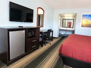 Habitación de hotel con cama, escritorio con silla, televisor, microondas, refrigerador, espejo y baño con lavamanos y espejo.
