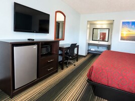 Habitación de hotel con cama, escritorio con silla, televisor, microondas, refrigerador, espejo y baño con lavamanos y espejo.
