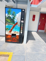 Una máquina expendedora con diseño de playa tropical, ubicada fuera de un edificio con puerta roja y un extintor en la pared.