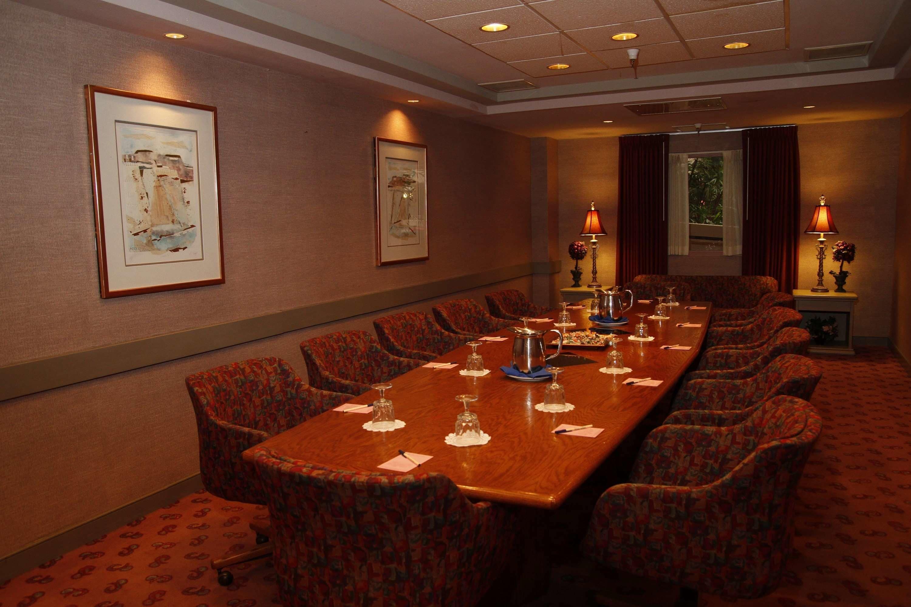 A conference room with a long table, patterned chairs, framed artwork on the walls, and a window with curtains.