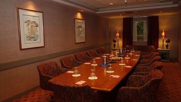 A conference room with a long table, patterned chairs, framed artwork on the walls, and a window with curtains.