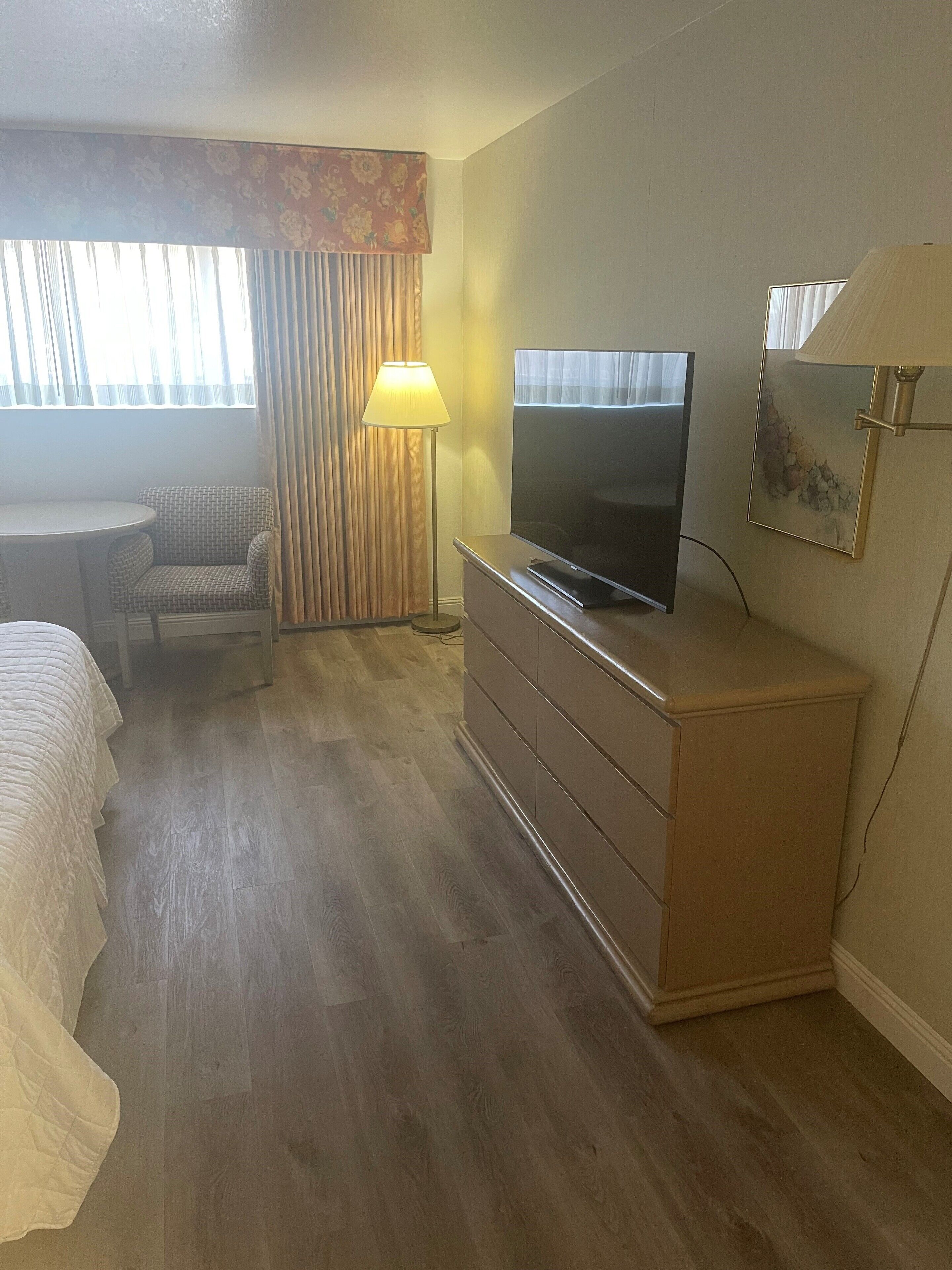 Ein Hotelzimmer mit Bett, Schreibtisch, Stuhl, Fernseher, Kommode und einem Fenster mit Vorhängen.