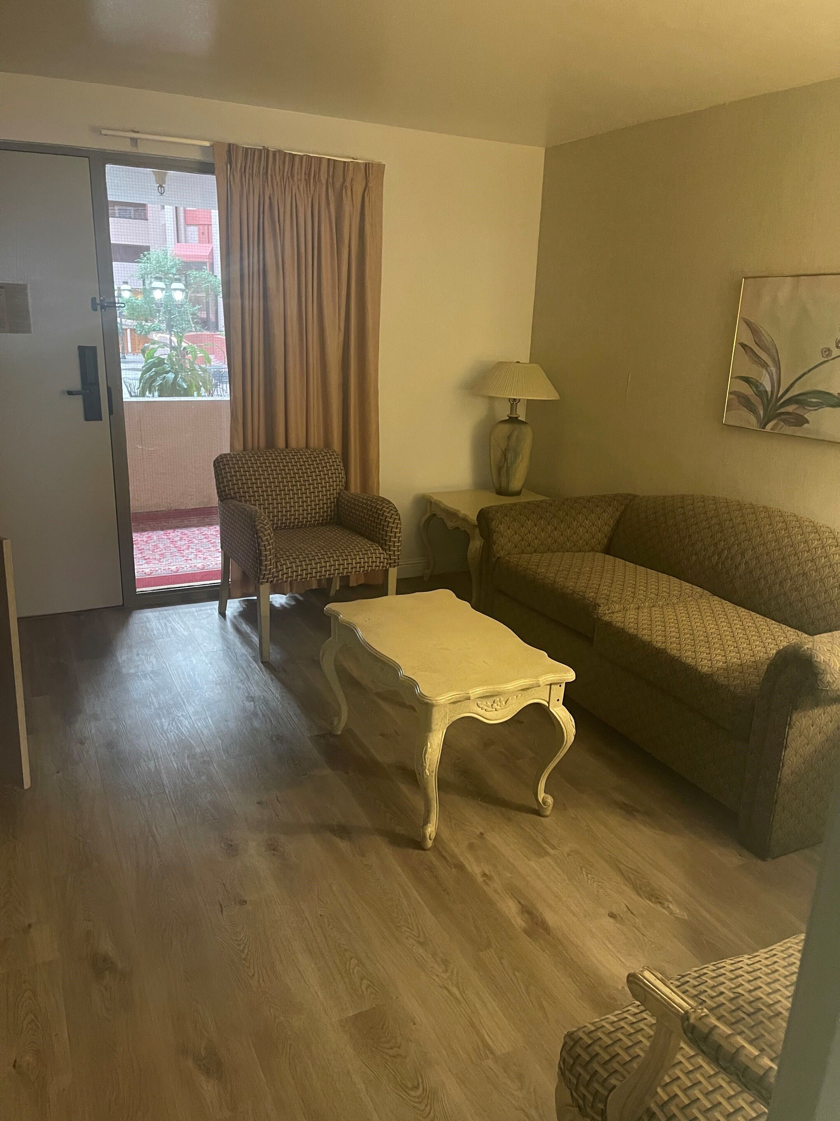 Ein Wohnzimmer mit Sofa, Sesseln und einem kleinen Tisch. Eine Tür führt in einen Außenbereich mit Blick auf Gebäude und eine Pflanze.