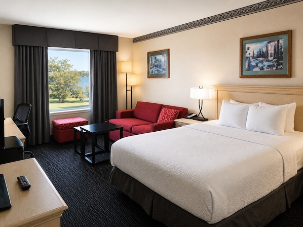 Una habitación de hotel con una cama grande, un sofá rojo, una mesita, un escritorio con silla, una lámpara y dos cuadros en la pared.