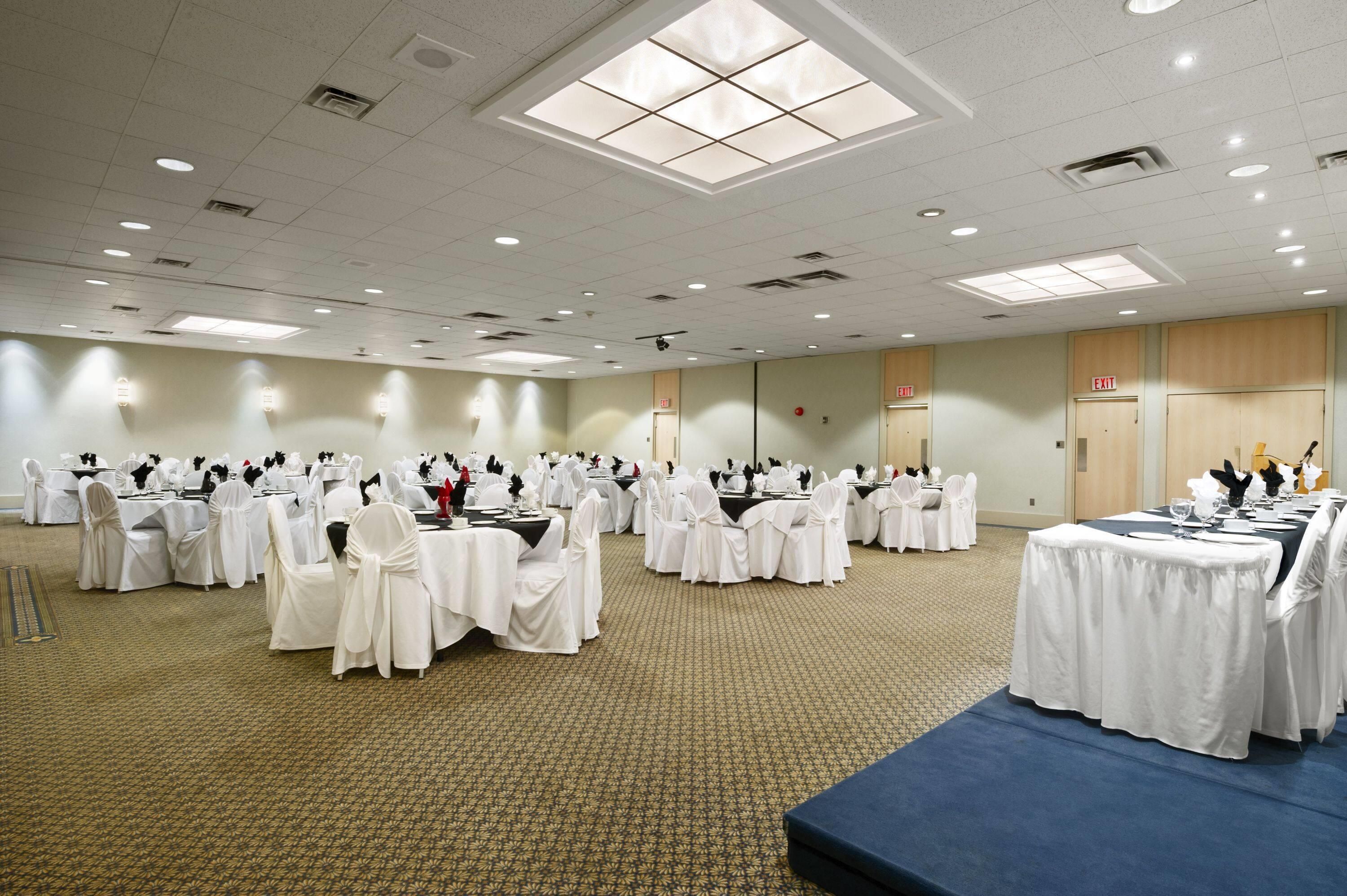 Un amplio salón de conferencias con varias mesas redondas, cada una cubierta con manteles blancos y sillas, y una zona de escenario con alfombra azul.