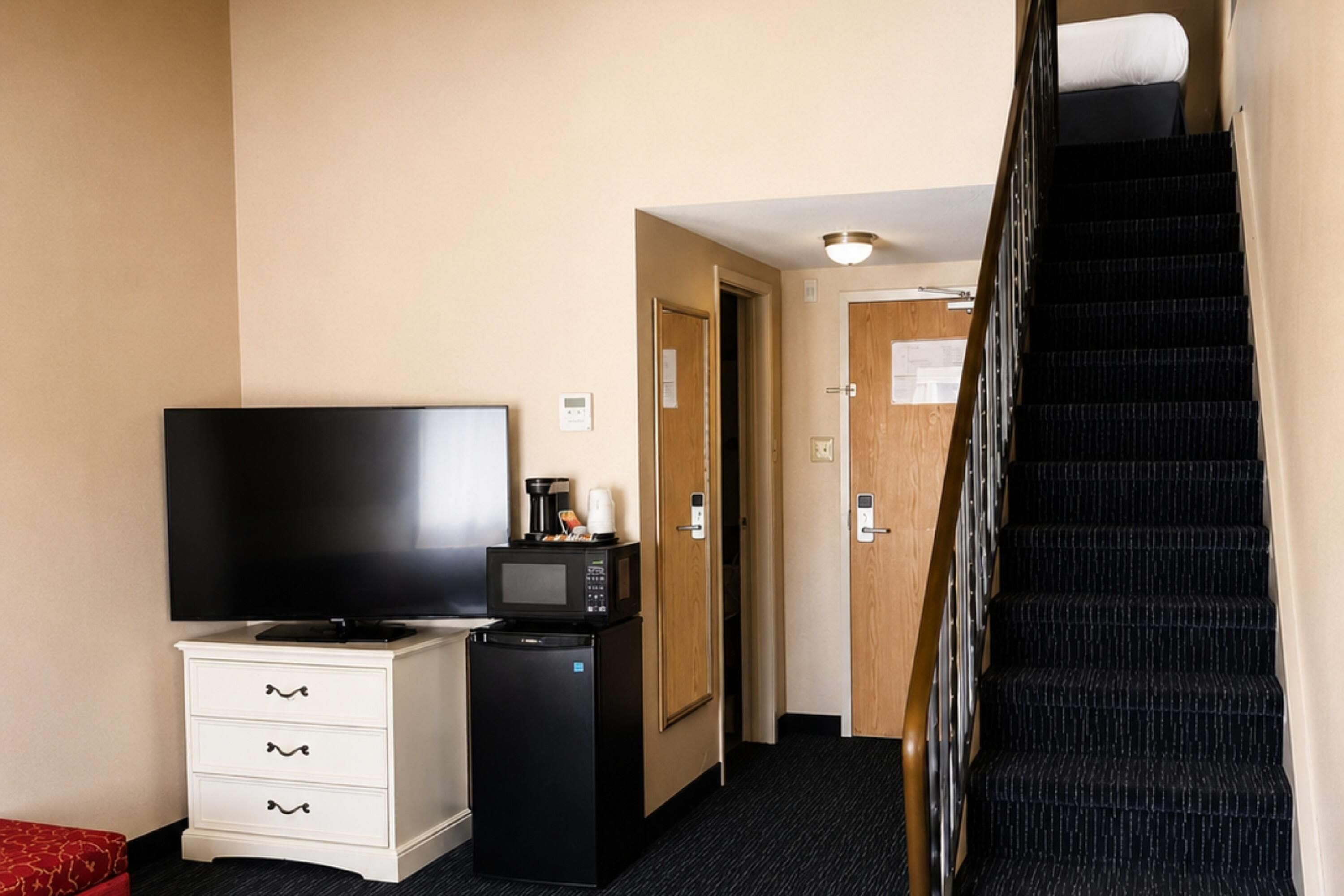 A hotel room with a flat-screen TV, a mini-fridge, a microwave, a small white cabinet, and a staircase leading to another room.