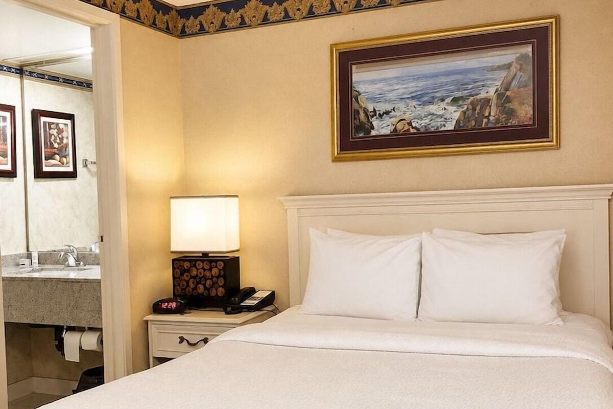Habitación de hotel con una cama grande, una mesita de noche con lámpara, un cuadro en la pared y un baño visible a través de una puerta abierta.