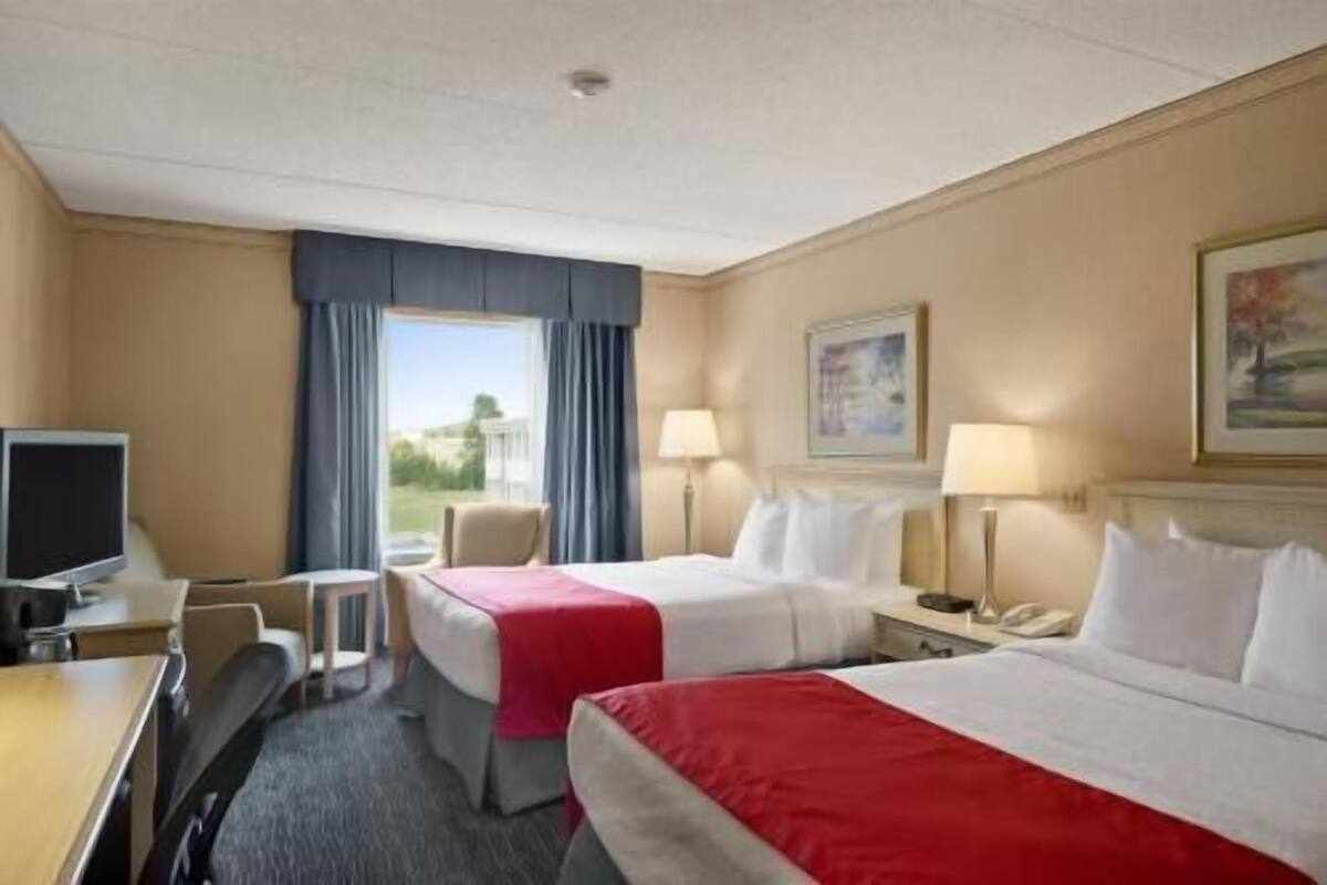 Habitación de hotel con dos camas, un escritorio con computadora, una silla, una ventana con cortinas y un cuadro en la pared.