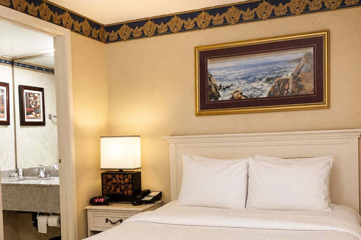 Habitación de hotel con cama, mesita de noche con lámpara, un cuadro en la pared y un baño visible a través de una puerta abierta.