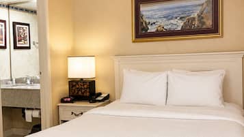 Une chambre d’hôtel avec un lit, une table de chevet avec une lampe, un tableau au mur et une salle de bain visible à travers une porte ouverte.