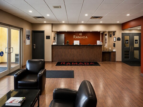 A reception area with a reception desk, two black leather chairs, a black reception mat, and a clock on the wall. - Ramada by Wyndham Trenton (Quinte West)
