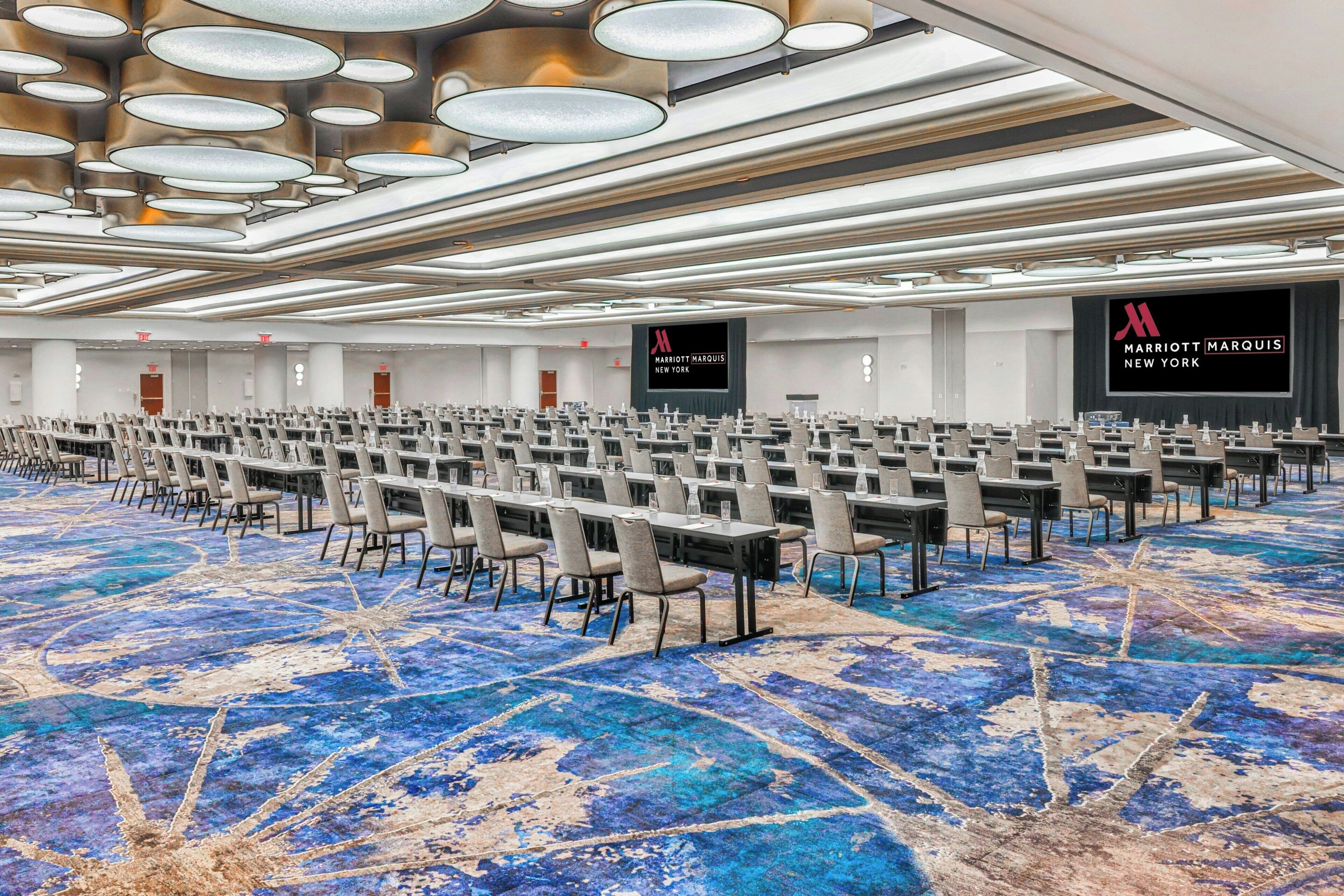 Une grande salle de conférence avec des rangées de tables et de chaises, un tapis à motifs bleus et un plafond orné de luminaires circulaires.