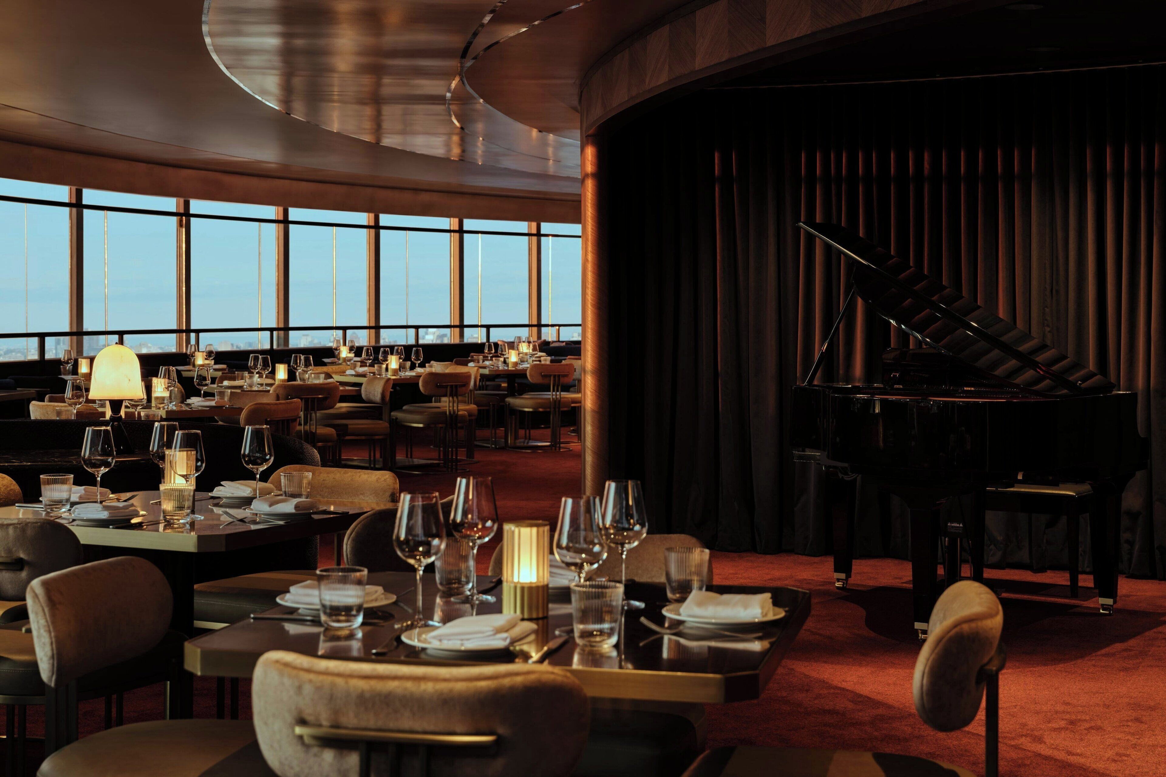 A spacious restaurant with tables set for dining, a grand piano, and large windows offering a view of the cityscape.