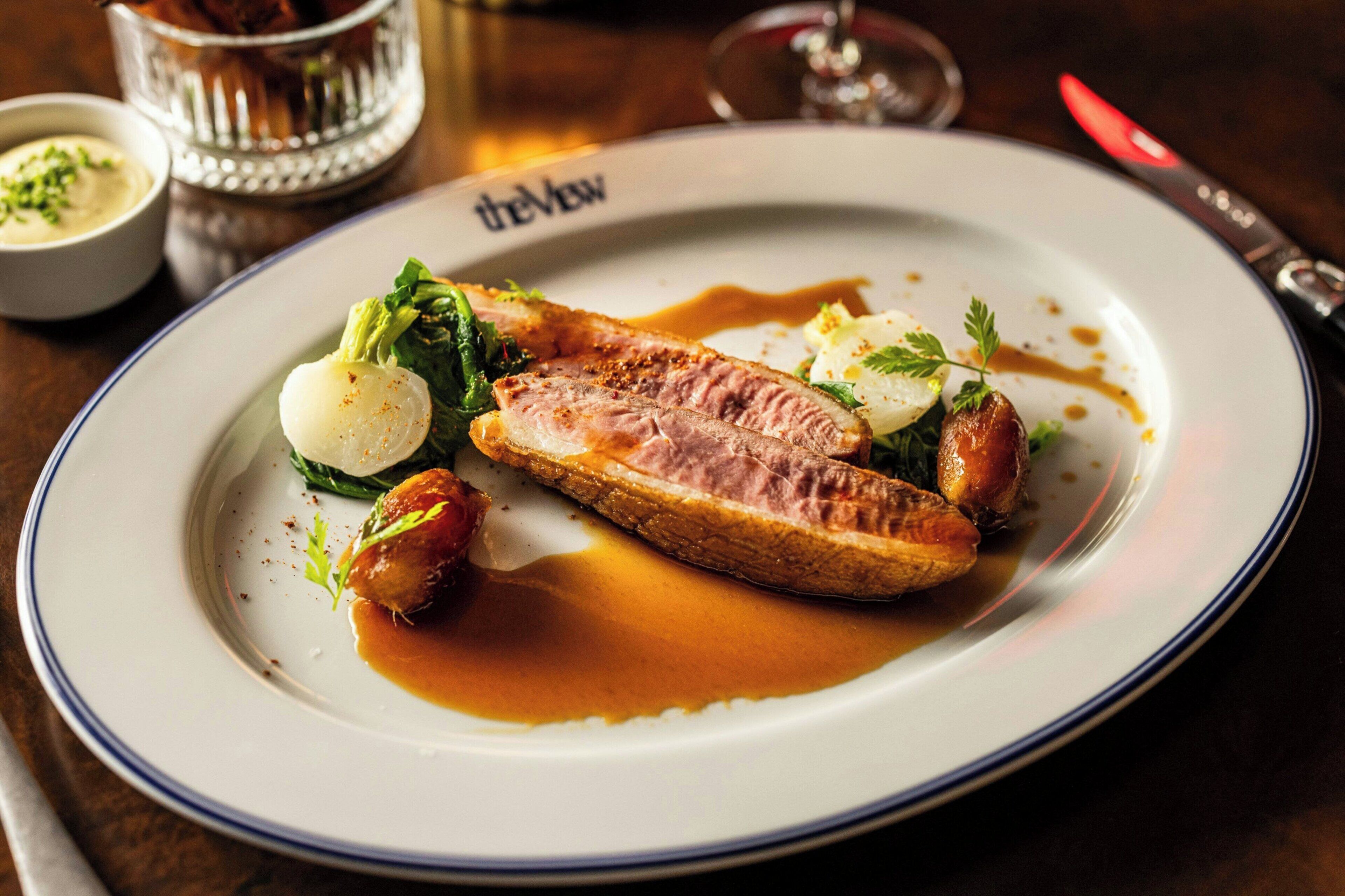Een bord eten met een stuk vlees, groenten en saus, op een wit bord met een blauwe rand.