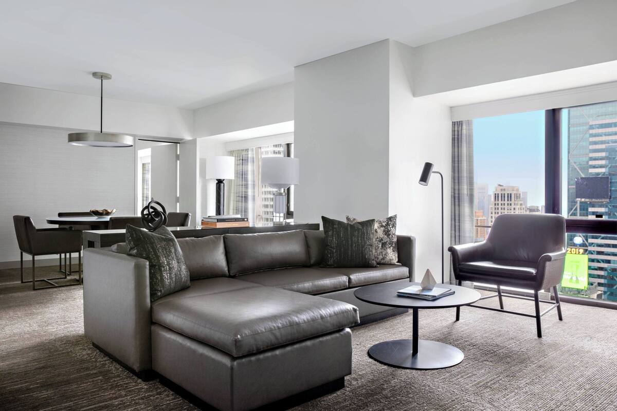 Een moderne hotelkamer met een leren bank, een rond bijzettafeltje, een eethoek met tafel en stoelen, en een groot raam met uitzicht over de stad.