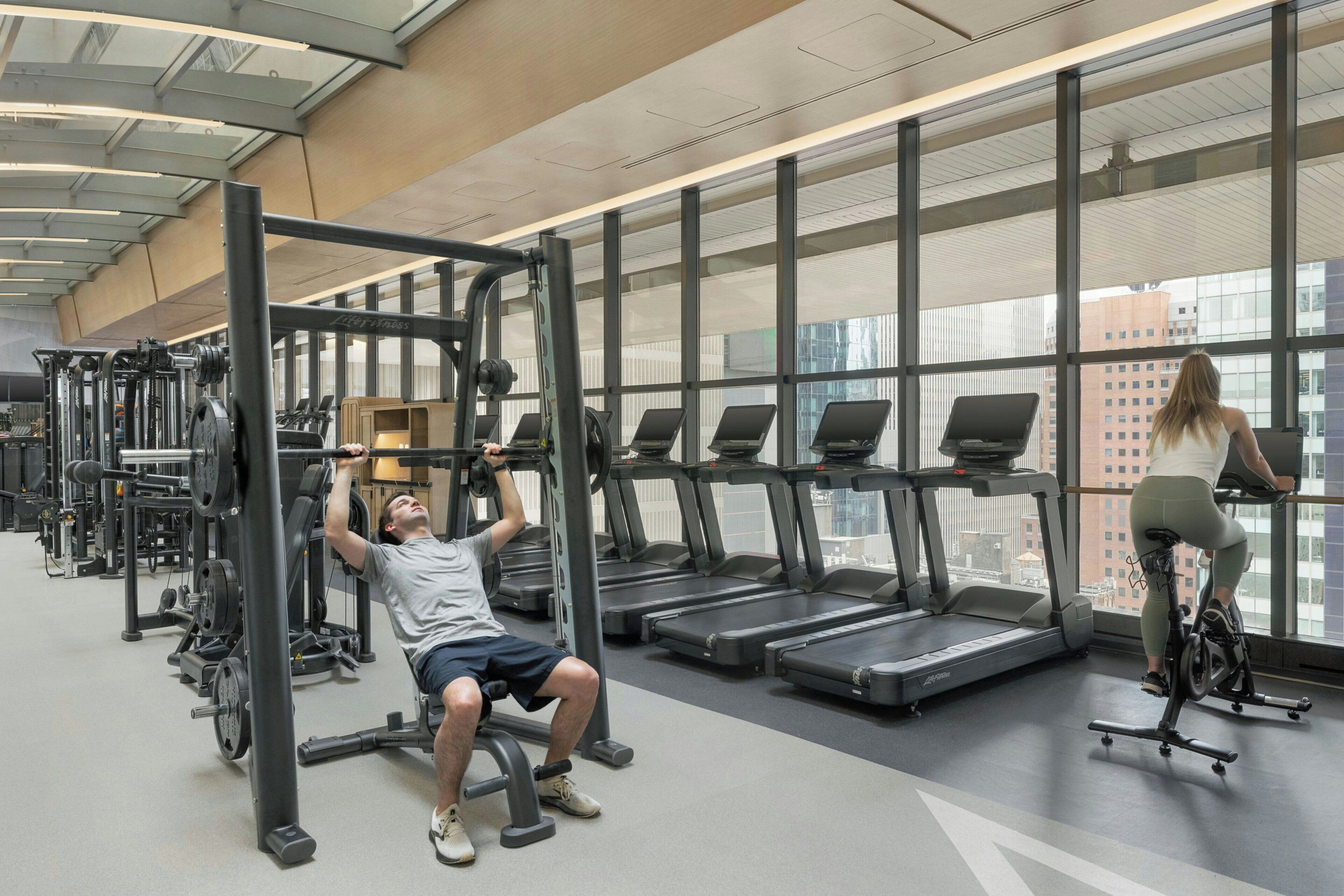 Un gimnasio con equipo de ejercicio, cintas de correr y vistas a edificios a través de amplios ventanales.