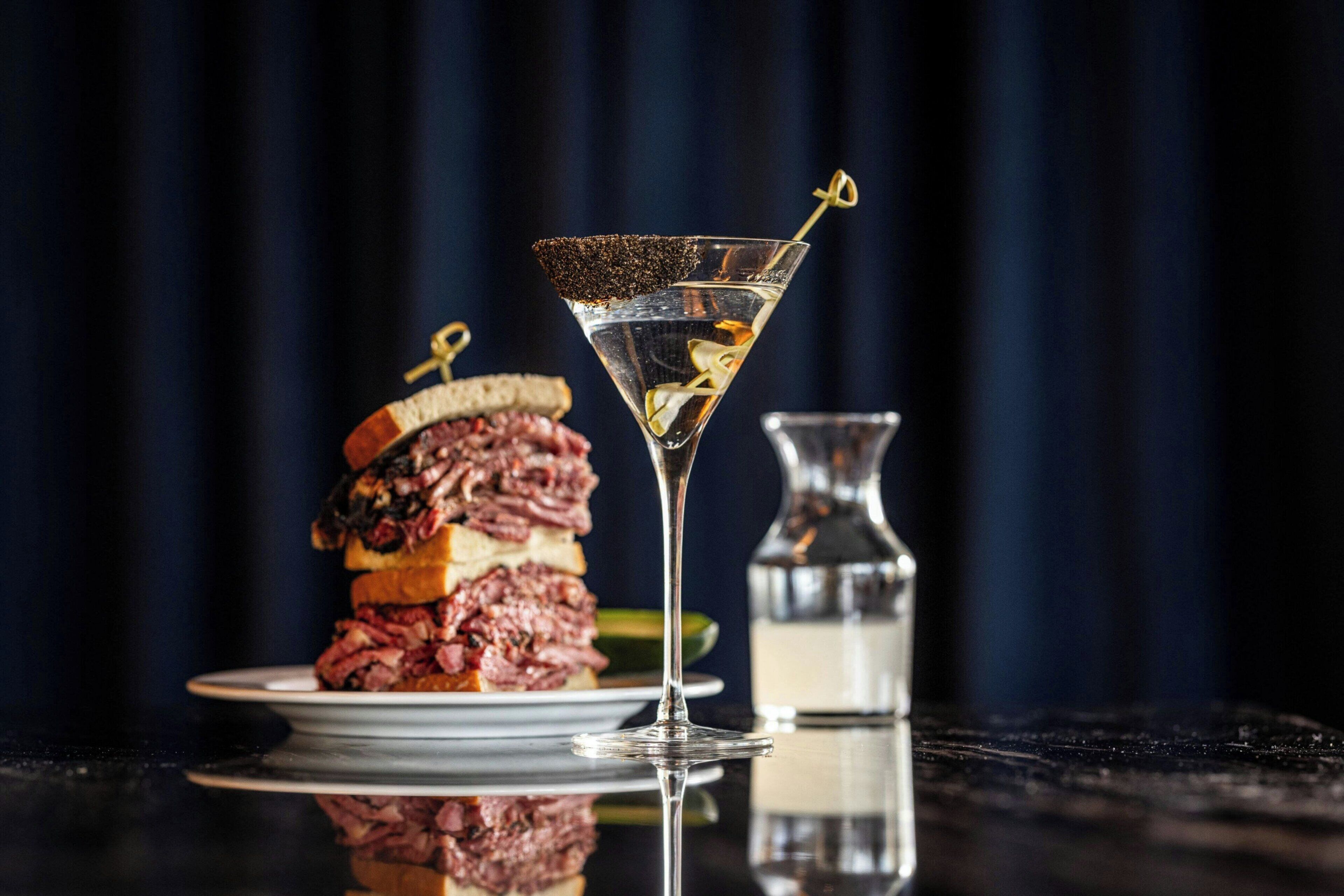 Una copa de martini con adorno sobre una superficie reflectante, un sándwich con decoración y una botella al fondo.