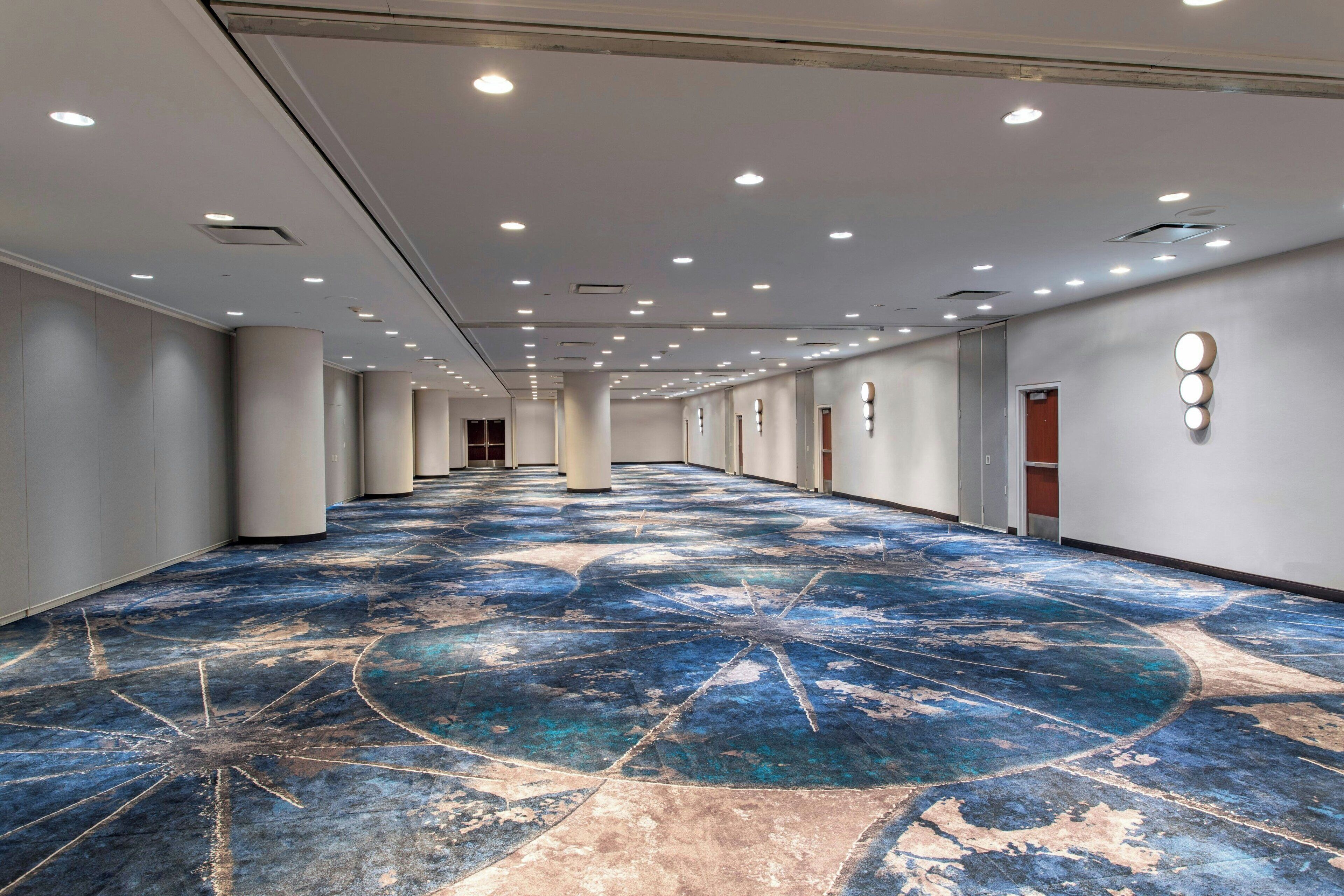 A spacious hotel corridor with a patterned floor, recessed lighting, and a series of doors along the walls.