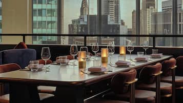 A dining area with a city view, featuring a long table set with glasses, plates, and candles.
