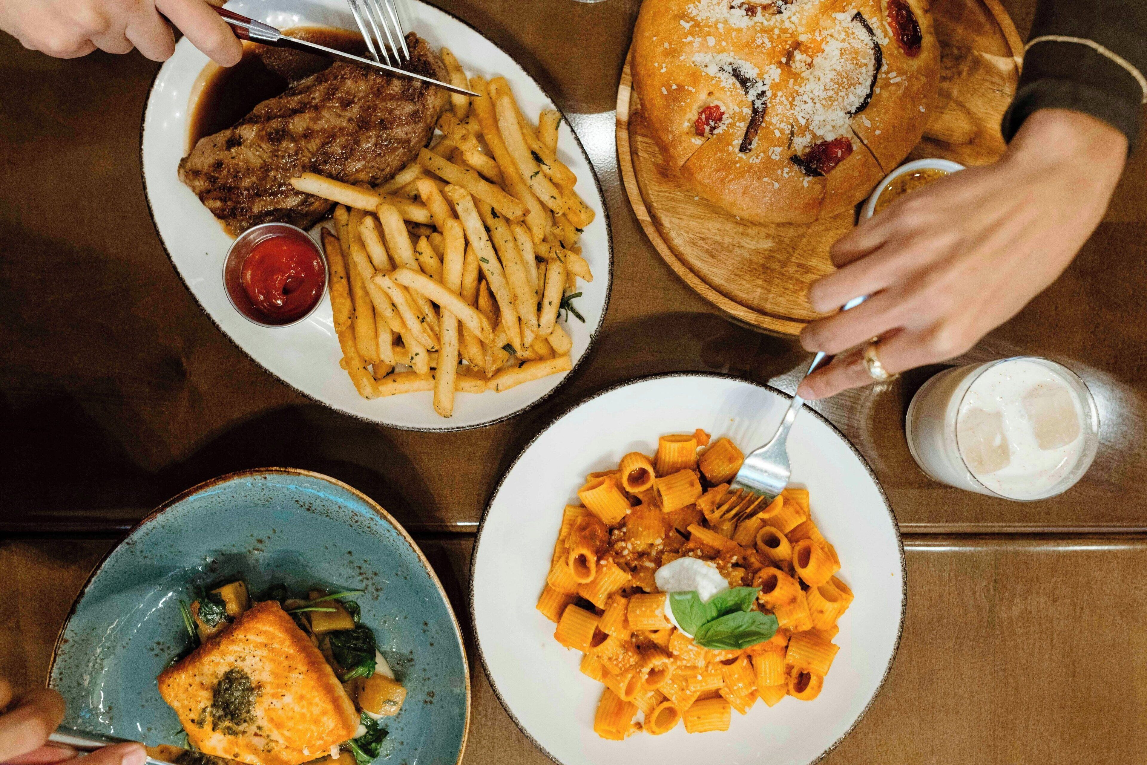 A table with three plates of food, including pasta, fries, and a meat dish, along with a bread roll and a glass of a clear beverage.