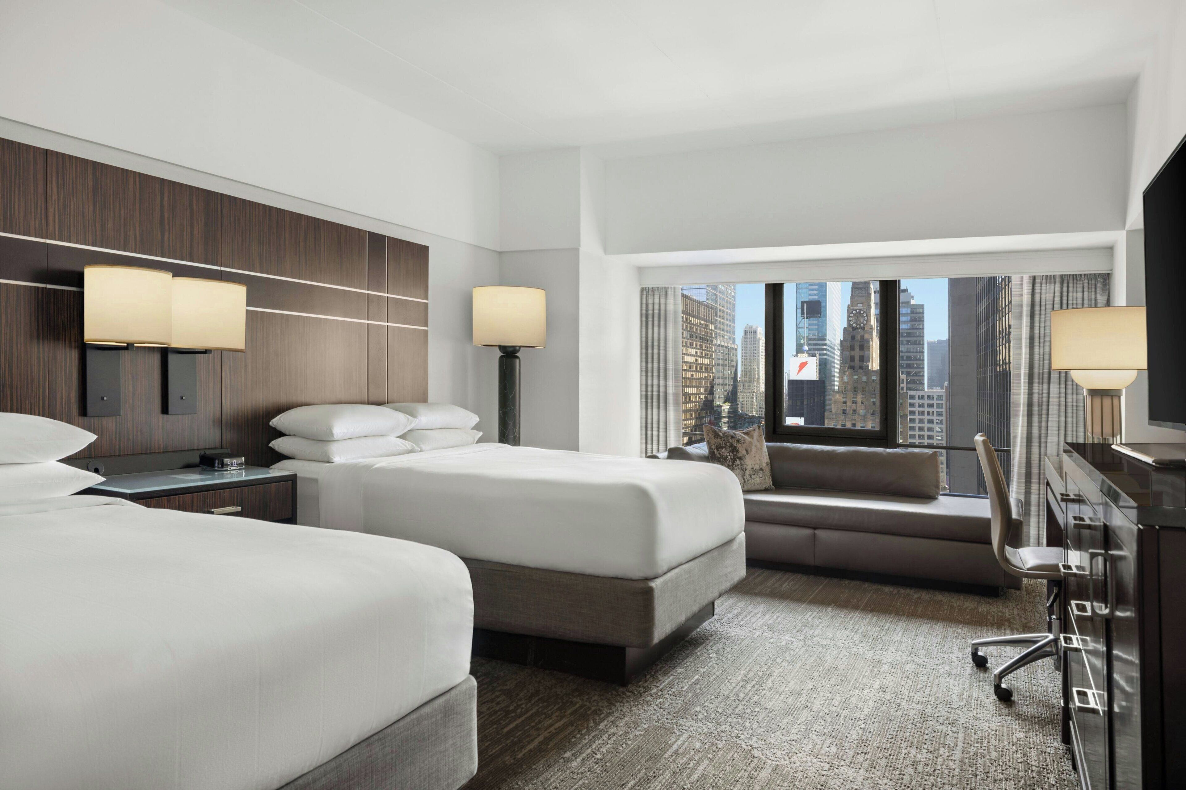 A hotel room with two beds, a sofa, a desk, and a large window with a city view.