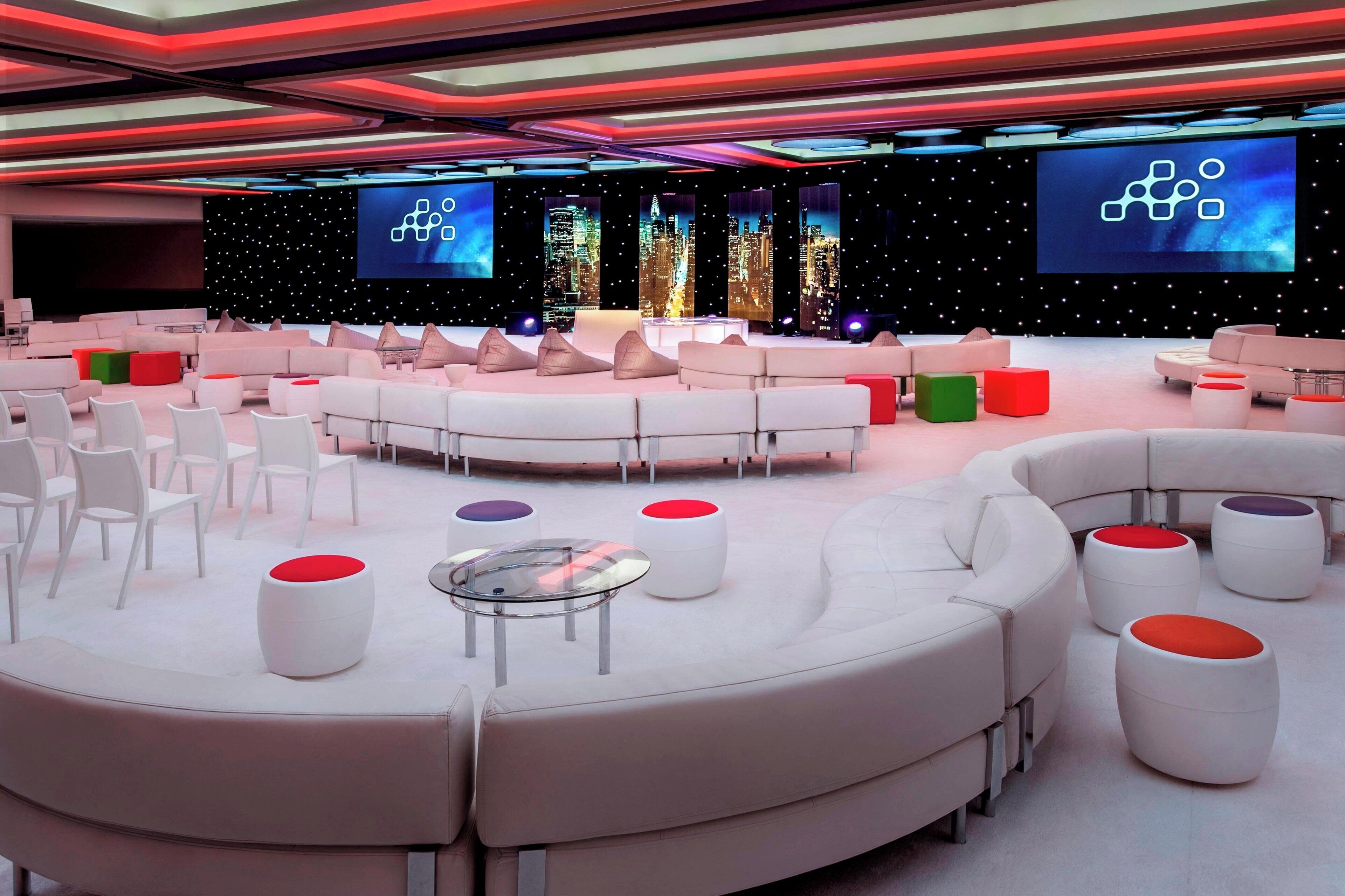 A modern lounge area with white sofas, red and green ottomans, and a large screen displaying a cityscape.