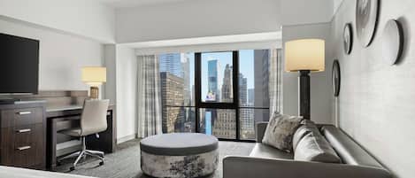 Habitación de hotel con cama, escritorio, silla, sofá, otomana redonda, televisor y ventana grande con vista a la ciudad.